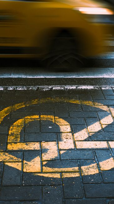 A close-up view of a parking space designated for disabled access, marked with a blue square and a white wheelchair symbol on cobblestone pavement. In the foreground, an orange traffic barrier with a