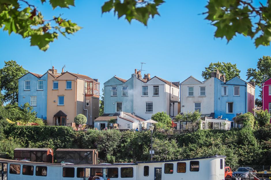 A row of multi-storey houses painted in various pastel colours, including blue, beige, light green, and pink, located on a hillside in a residential neighbourhood. The houses have small front gardens